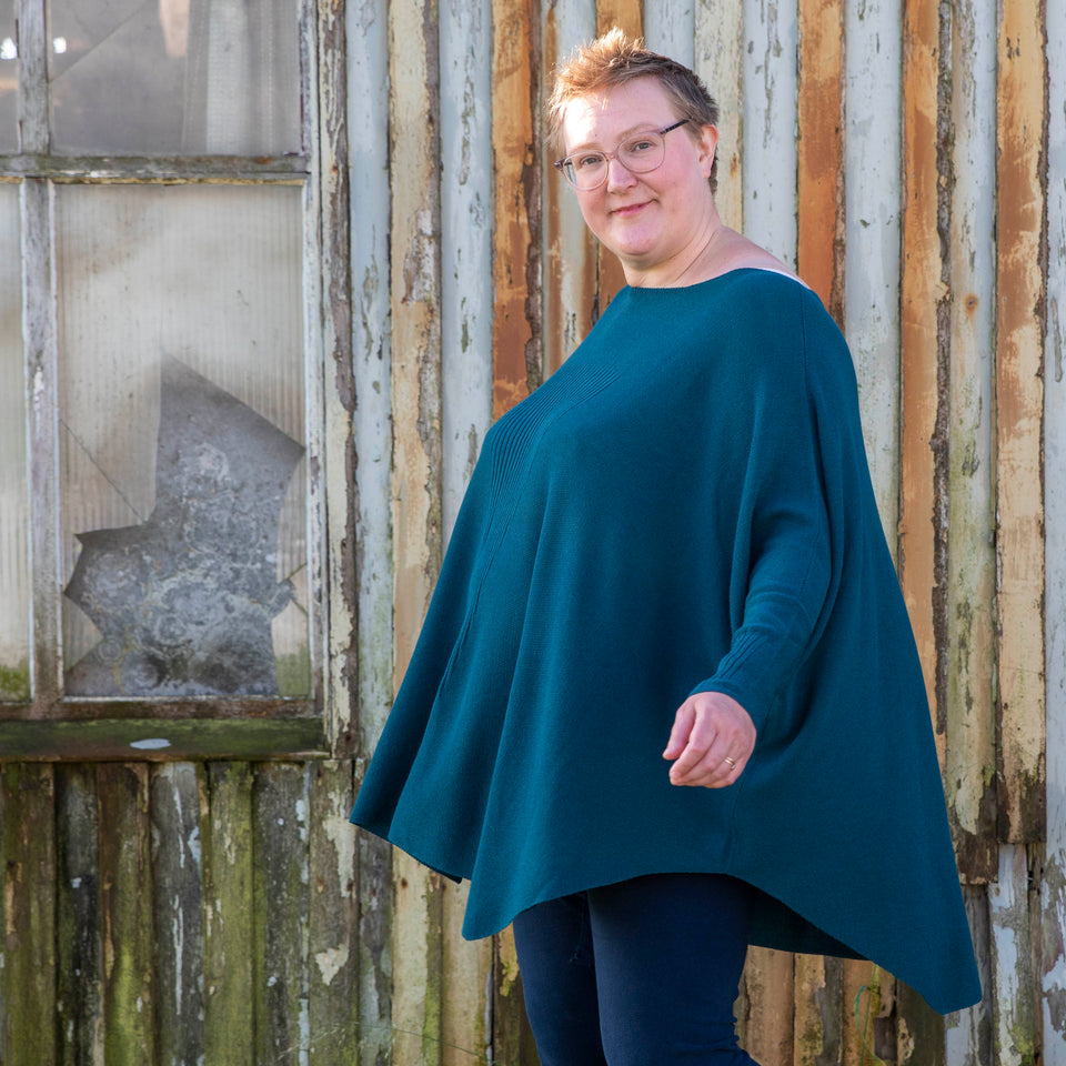 A white woman with short fair hair walks in front of the Hoswick Gospel Hall - and old wooden chapel that is now worn by weather. She is wearing clear framed spectacles and an airy dark teal, lightweight jumper. The body of the jumper is full and loose and it has dolman sleeves. The jumper swings out as she moves. Underneath she has navy leggings. She turns her head slightly and looks towards the camera.