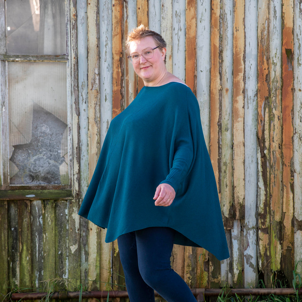 A white woman with short fair hair walks in front of the Hoswick Gospel Hall - and old wooden chapel that is now worn by weather. She is wearing clear framed spectacles and an airy dark teal, lightweight jumper. The body of the jumper is full and loose and it has dolman sleeves. The jumper swings out as she moves. Underneath she has navy leggings. She turns her head slightly and looks towards the camera.