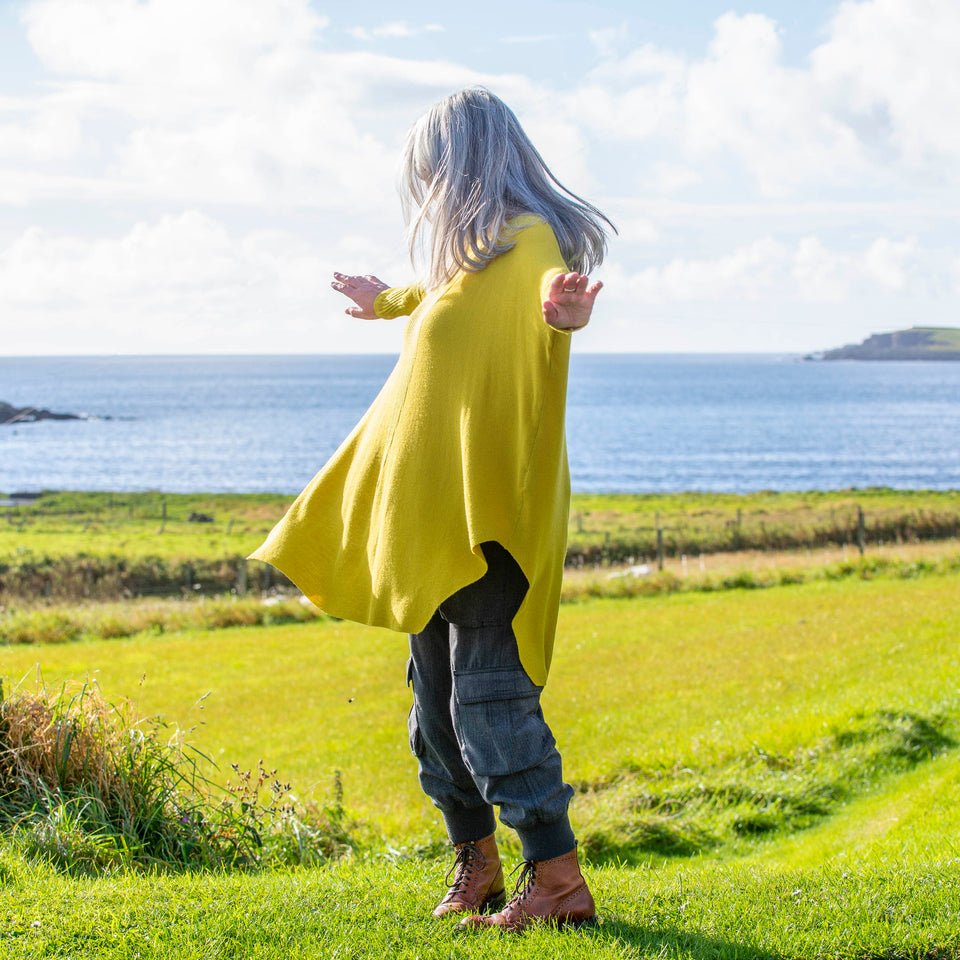 A white woman with long, straight grey hair stands at the top of a field in Shetland overlooking sea. She stands side on to the camera and spins with her arms out. She wears a lightweight, long yellow jumper over charcoal combat trousers and brown lace up boots