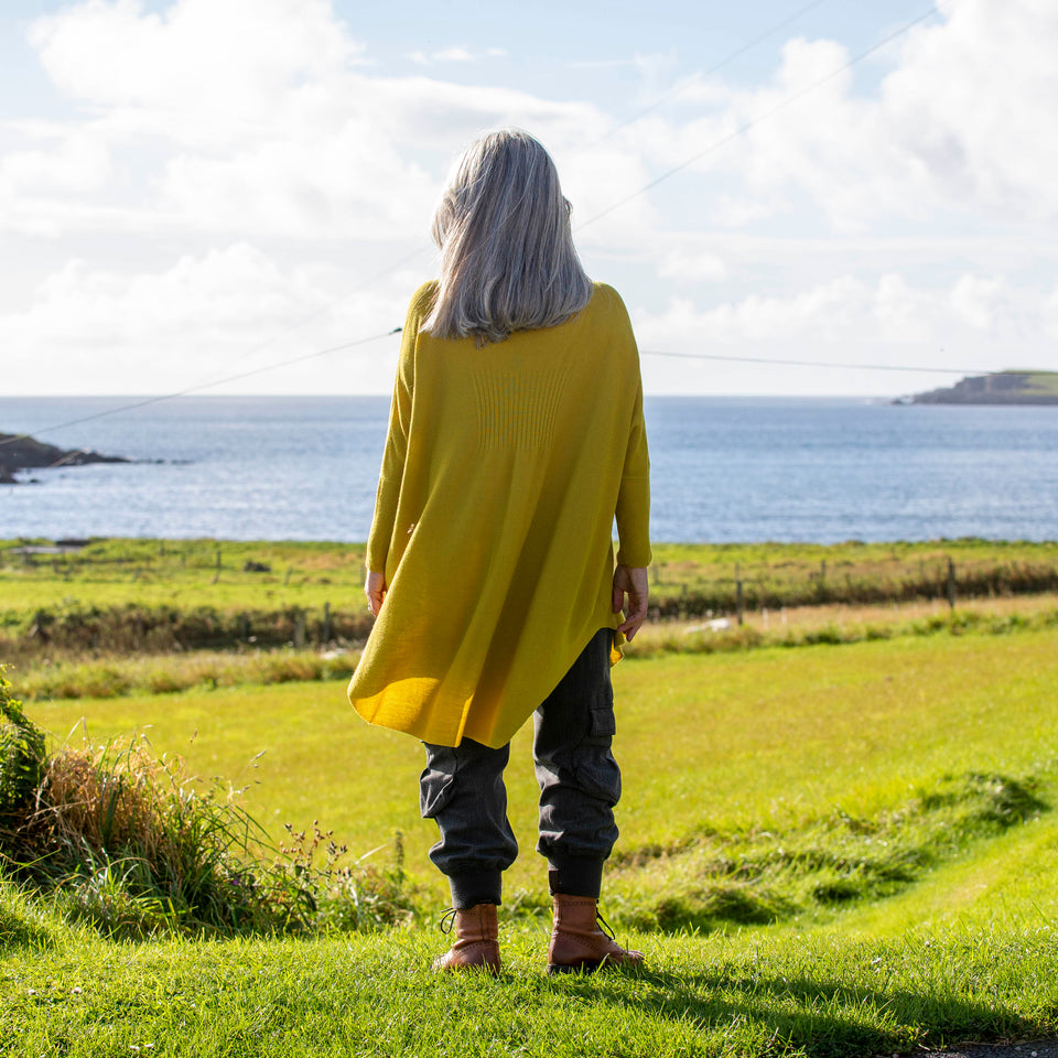 A white woman with long, straight grey hair stands at the top of a field in Shetland overlooking the sea. She wears a lightweight, long yellow jumper over charcoal combat trousers and brown lace up boots