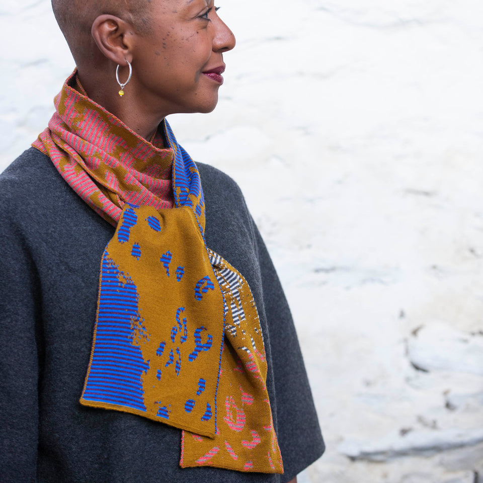 A black woman with a shaved head looks to her left. She is wearing a conntemporary Scottish finely knitted scarf in a pattern of stripes and mottled areas. Pinks, ochres, blues, mustards interchange. She also wears a charcoal woollen dress.