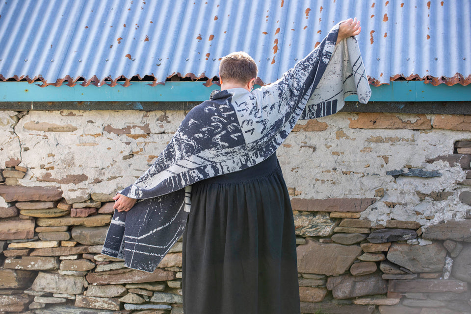 A white woman with short fair hair stands in front of an old stone building with a corrugated tin roof in Hoswick, Shetland. She wears glasses with clear frames and a short sleeved black heavy linen dress with full skirt. She has her back to the camera and stretches out her arms to show a large shawl/wrap in a contemporary design in charcoal and off white abstract design. Photograph taken in Hoswick, Shetland, Scotland.