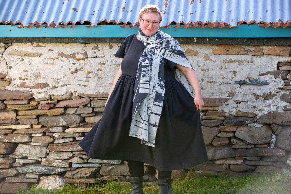 A white woman with short fair hair stands in front of an old stone building with a corrugated tin roof in Hoswick, Shetland. She wears glasses with clear frames and a short sleeved black heavy linen dress with full skirt. She wears a large shawl/wrap in a contemporary design in charcoal and off white abstract design. Photograph taken in Hoswick, Shetland, Scotland.