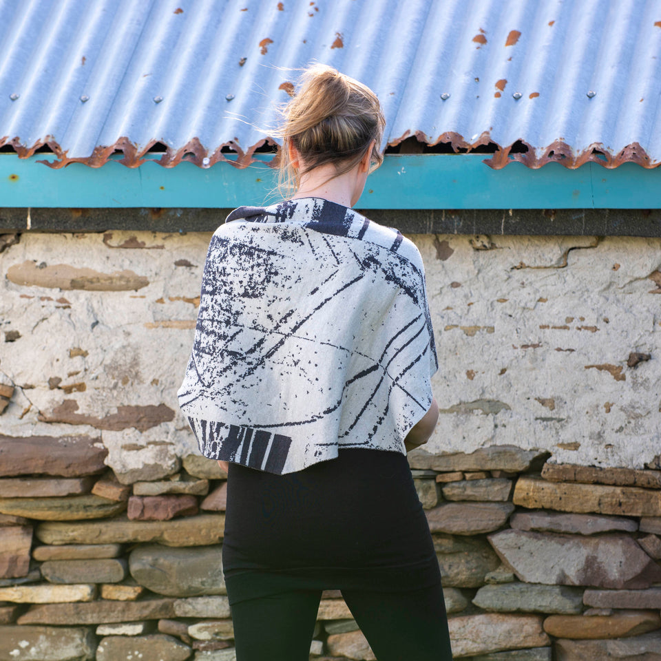 A white woman with light brown hair tied back stands in front of a stone shed with a corrugated tin roof. She is wearing a contempoarary scottish knit cape in charcoal grey and off white over a black vest and leggings. View from the back