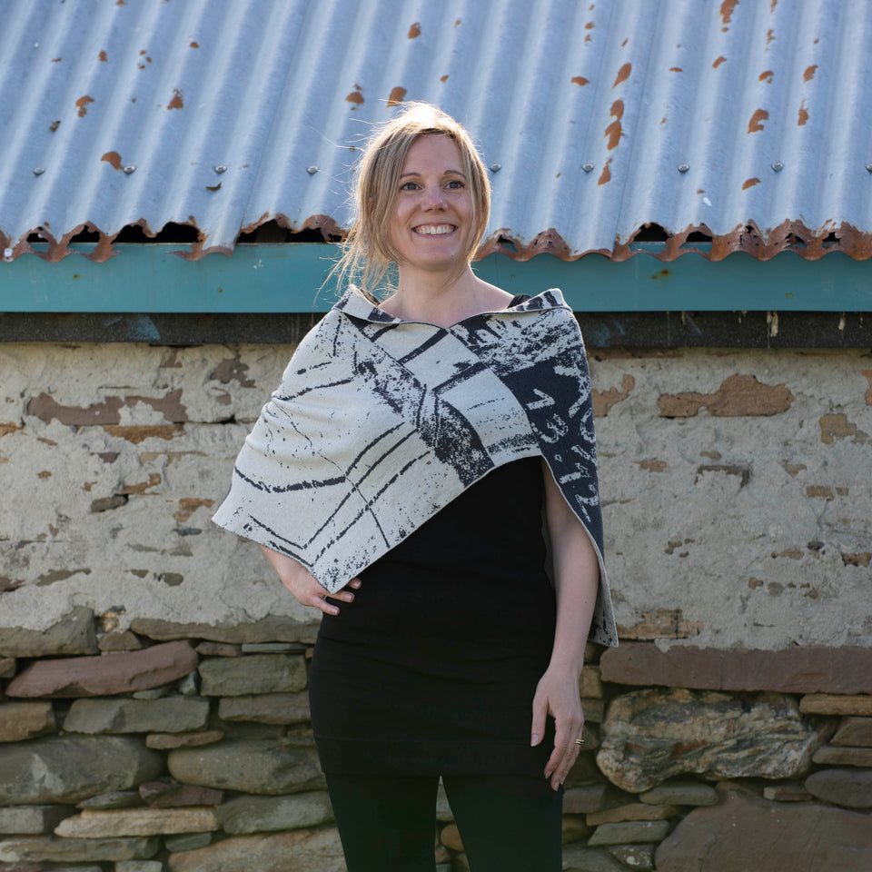 A white woman with light brown hair tied back stands in front of a stone shed with a corrugated tin roof. She is wearing a contempoarary scottish knit cape in charcoal grey and off white over a black vest and leggings.