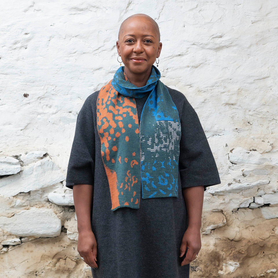 A black woman with a shaved head stands in a rustic, whitewashed stone building in Shetland. She wears a charcoal woollen dress with elbow-length sleeves. Over this she wears a finely knitted Scottish made scarf in a stripey and mottled pattern. Greens, blues and oranges interchange. She looks at the camera and smiles.