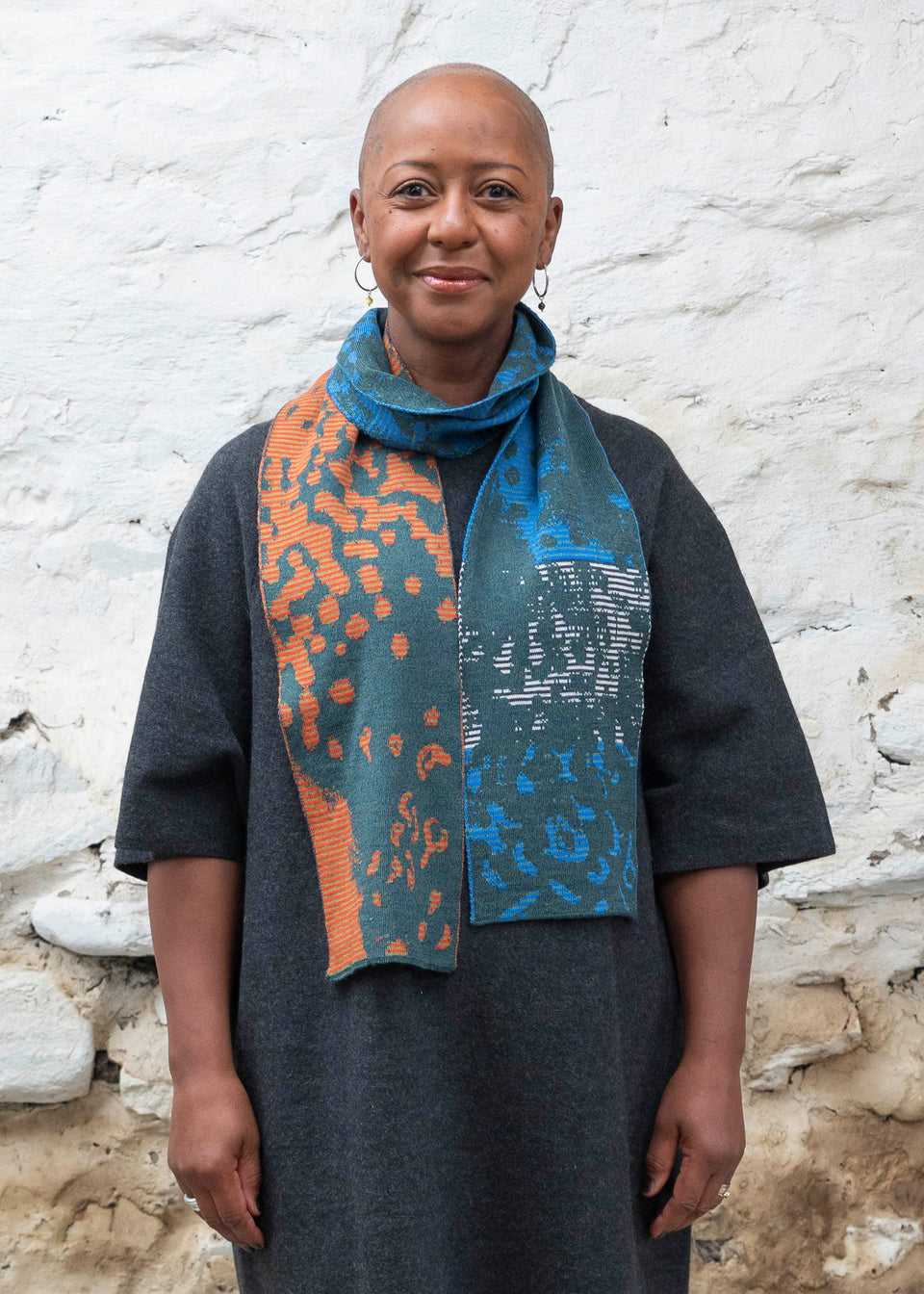 A black woman with a shaved head stands in a rustic, whitewashed stone building in Shetland. She wears a charcoal woollen dress with elbow-length sleeves. Over this she wears a finely knitted Scottish made scarf in a stripey and mottled pattern. Greens, blues and oranges interchange. She looks at the camera and smiles.