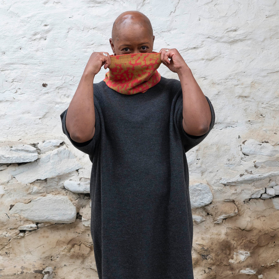 A black woman with a shaved head wears a finely knitted circle scarf in bright coral and gold merino wool, over a charcoal grey dress. She is standing in a rustic building with whitewashed walls in Shetland.