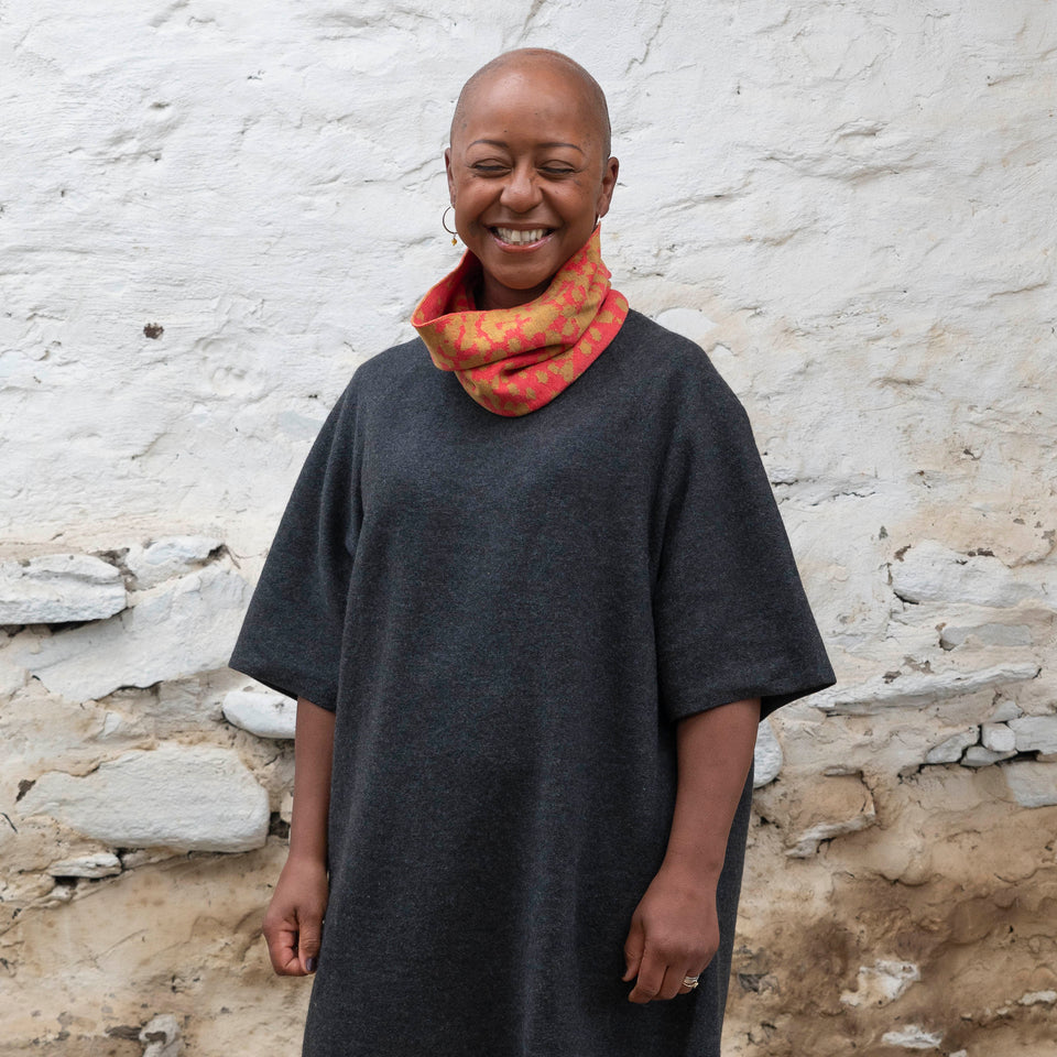 A black woman with a shaved head wears a finely knitted circle scarf in bright coral and gold merino wool, over a charcoal grey dress. She is standing in a rustic building with whitewashed walls in Shetland.