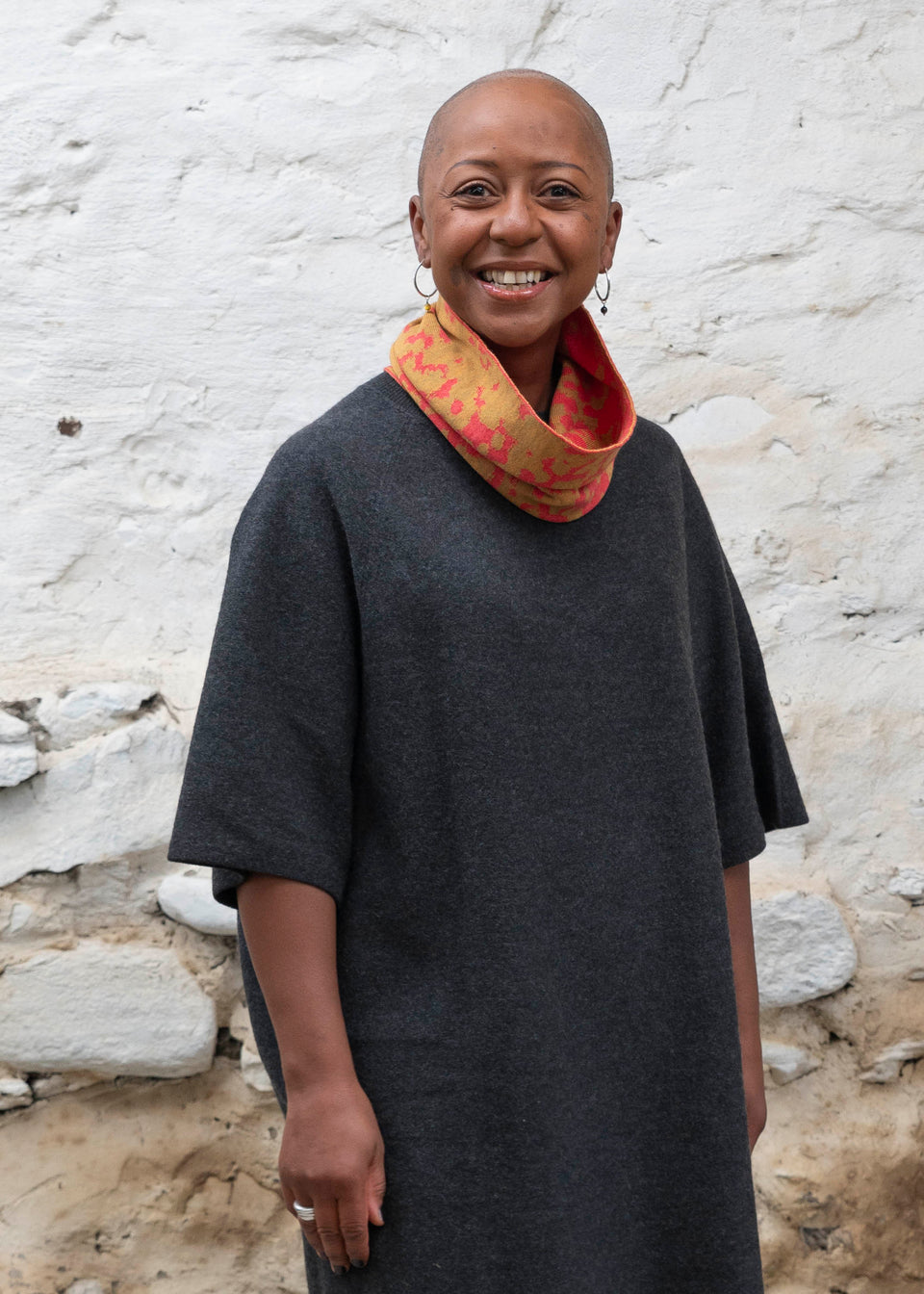 A black woman with a shaved head wears a finely knitted circle scarf in bright coral and gold merino wool, over a charcoal grey dress. She is standing in a rustic building with whitewashed walls in Shetland.