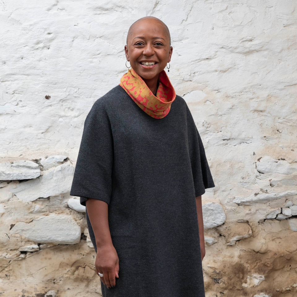 A black woman with a shaved head wears a finely knitted circle scarf in bright coral and gold merino wool, over a charcoal grey dress. She is standing in a rustic building with whitewashed walls in Shetland.