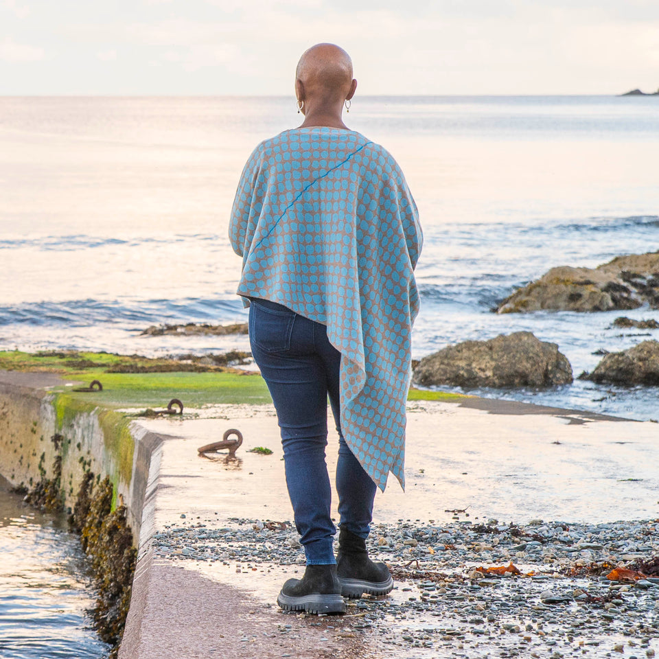 On the concrete pier at Hoswick, Shetland, Jeanette Sloan wears an asymmetric, contemporary sweater with long ribbedcuffs and an all-over ireegular pebble pattern in soft blues and greys. The statement sweater is worn over dark indigo jeans and chunky black boots. The day is calm and the soft copper light reflects on the sea.