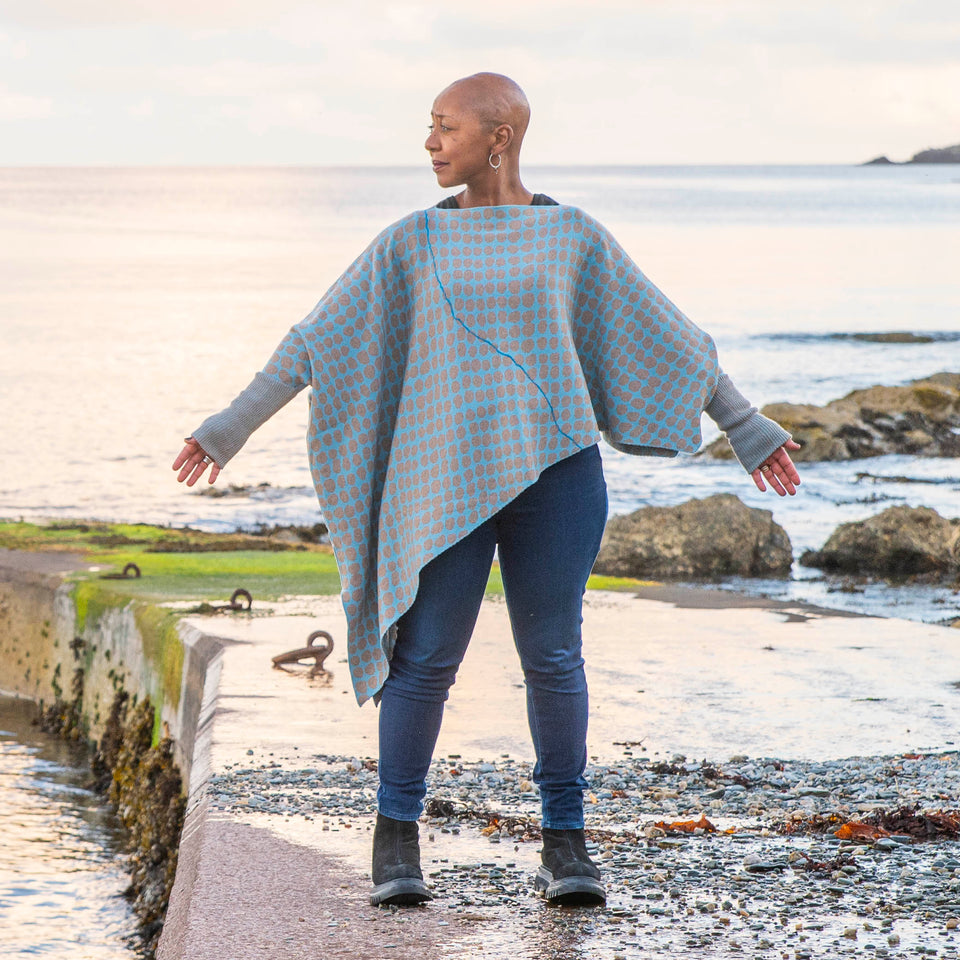 On the concrete pier at Hoswick, Shetland, Jeanette Sloan wears an asymmetric, contemporary sweater with long ribbedcuffs and an all-over ireegular pebble pattern in soft blues and greys. The statement sweater is worn over dark indigo jeans and chunky black boots. The day is calm and the soft copper light reflects on the sea.