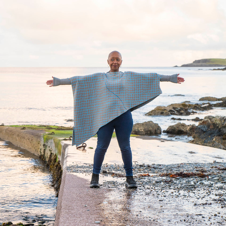 On the concrete pier at Hoswick, Shetland, Jeanette Sloan wears an asymmetric, contemporary sweater with long ribbedcuffs and an all-over ireegular pebble pattern in soft blues and greys. The statement sweater is worn over dark indigo jeans and chunky black boots. The day is calm and the soft copper light reflects on the sea.
