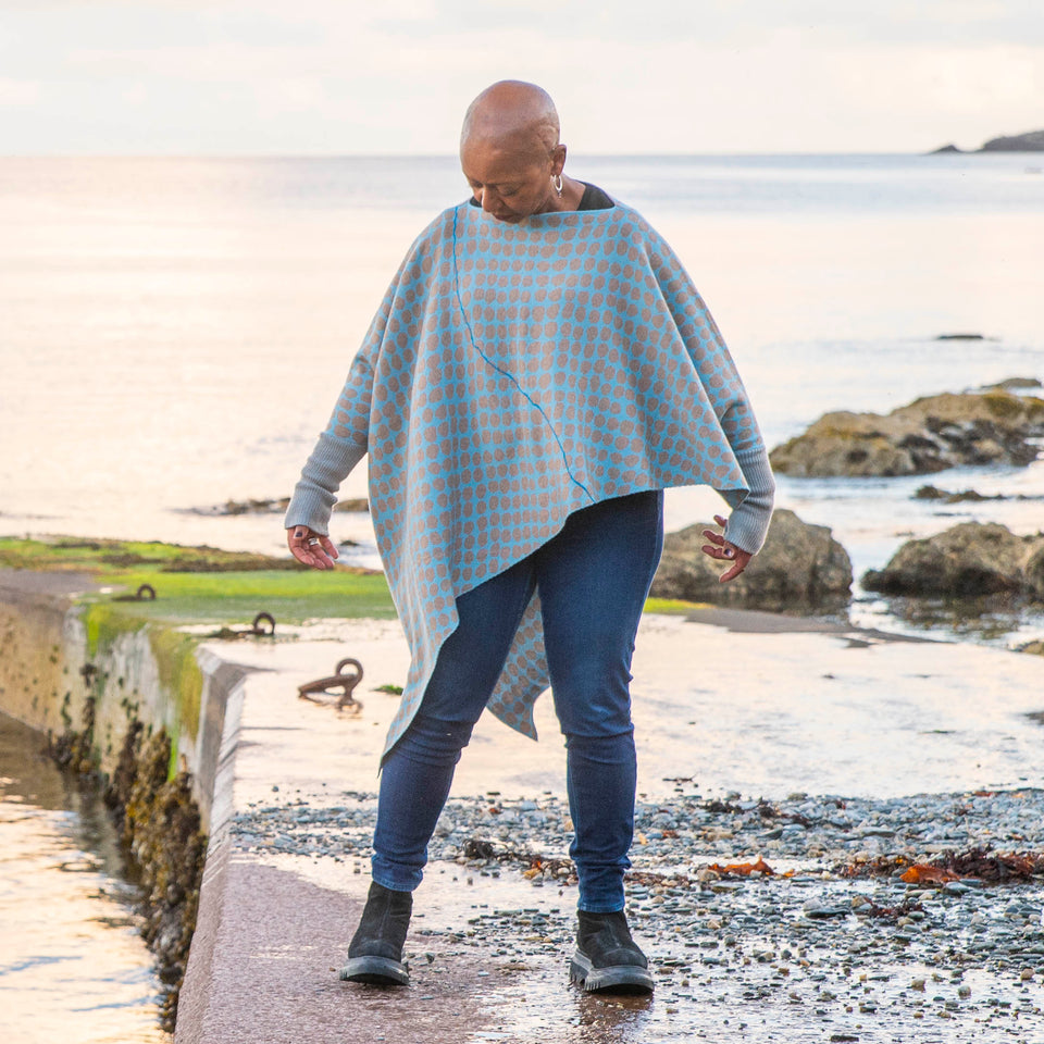 On the concrete pier at Hoswick, Shetland, Jeanette Sloan wears an asymmetric, contemporary sweater with long ribbedcuffs and an all-over ireegular pebble pattern in soft blues and greys. The statement sweater is worn over dark indigo jeans and chunky black boots. The day is calm and the soft copper light reflects on the sea.