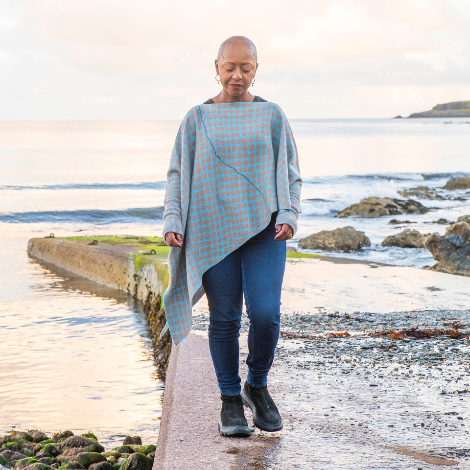 On the concrete pier at Hoswick, Shetland, Jeanette Sloan wears an asymmetric, contemporary sweater with long ribbedcuffs and an all-over ireegular pebble pattern in soft blues and greys. The statement sweater is worn over dark indigo jeans and chunky black boots. The day is calm and the soft copper light reflects on the sea.