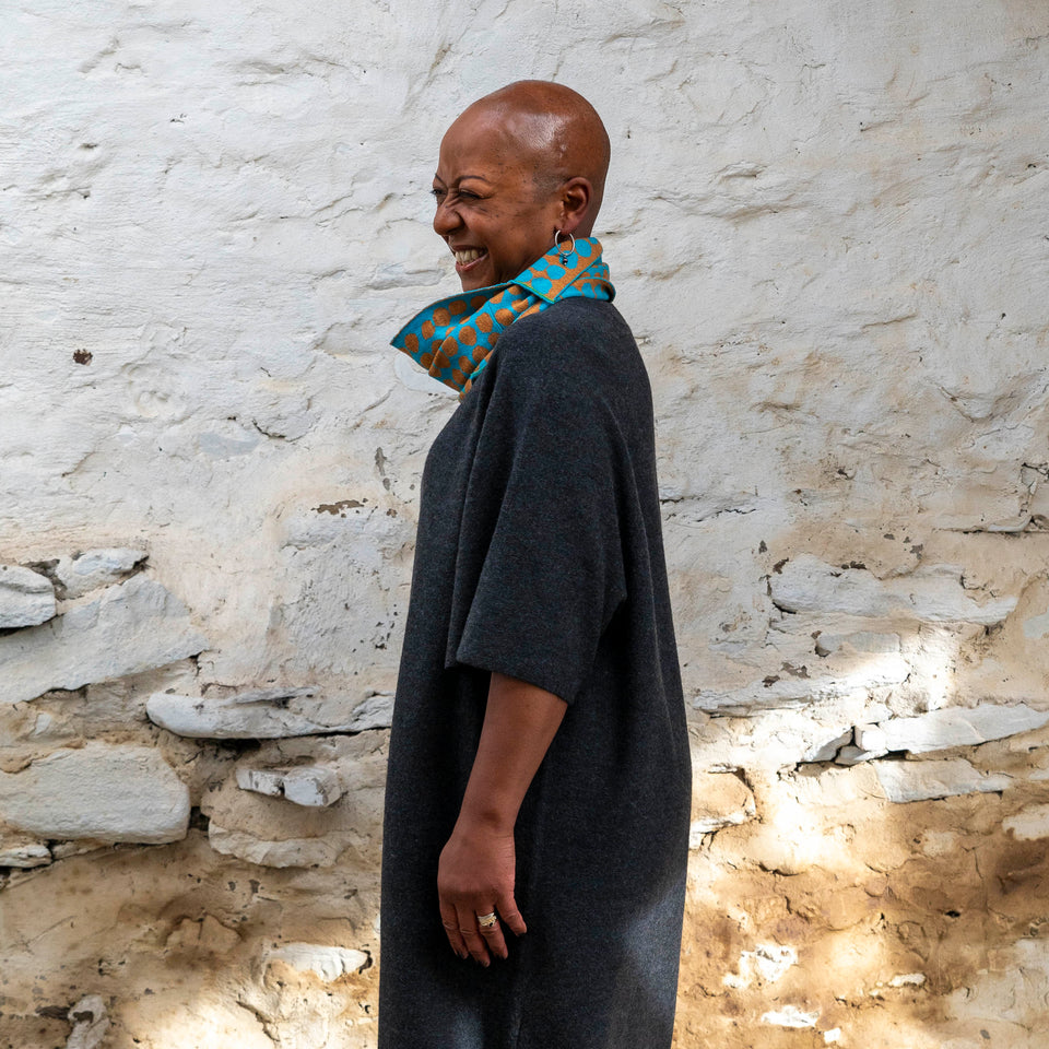 Merino circle scarf - a black woman with shaved head stands in a rustic stone building in Hoswick, shetland. She is wearing a charcoal wool dress with elbow length sleeves . Around her neck is a merino  loop scarf in ochre and turquoise.