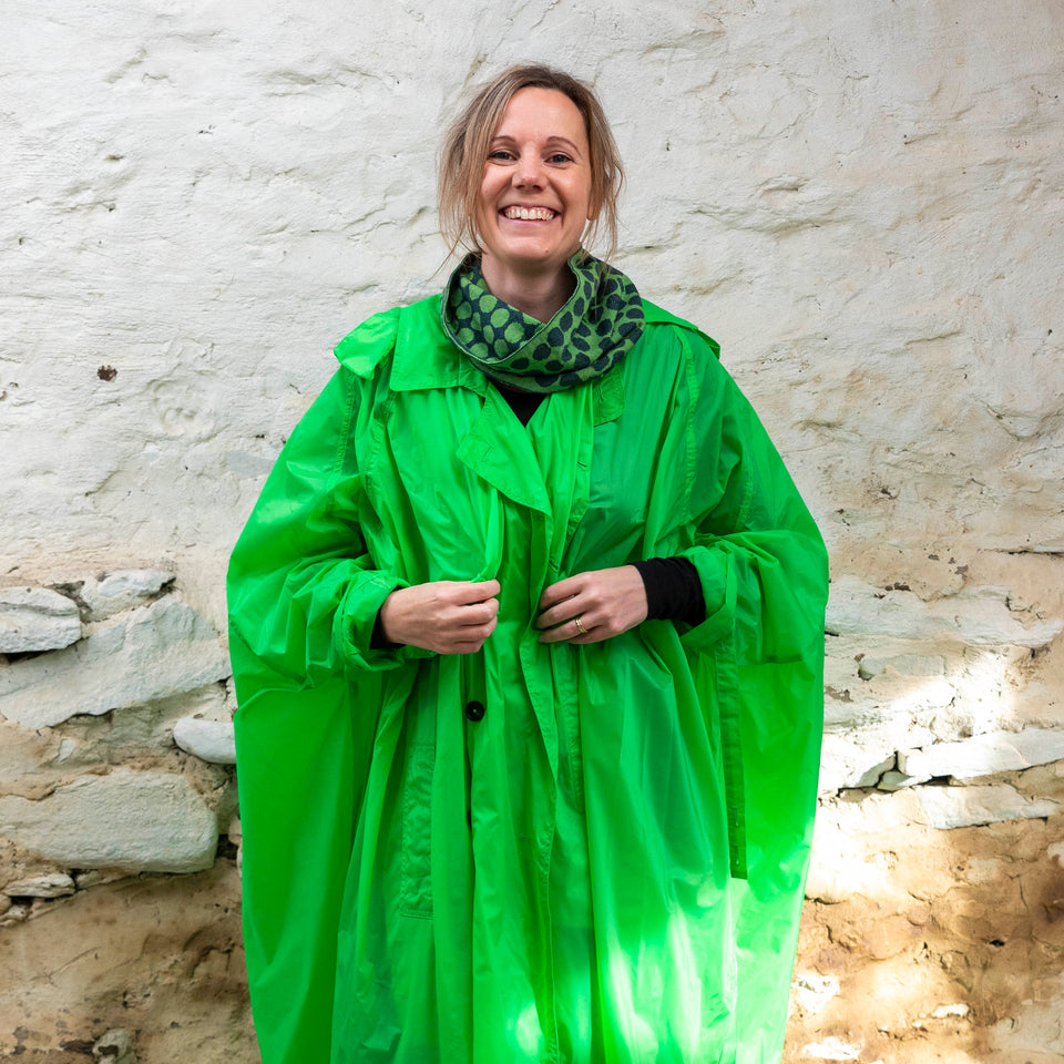 Merino loop scarf - A white woman with fair hair tied back stands in a rustic stone building in Hoswick, Shetland Islands, Scotland. She is wearing a green light weight rain coat  over jeans and a circle scarf with irregular dot design in green and navy. 