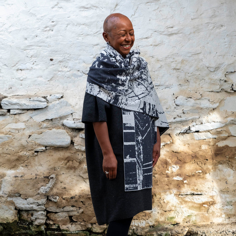 A black woman with a shaved head stands in a rustic stone building with whitewashed walls in Hoswick, Shetland. She wears black tights, a charcoal and off white shawl is wrapped around her.. She shuts her eyes and laughs.