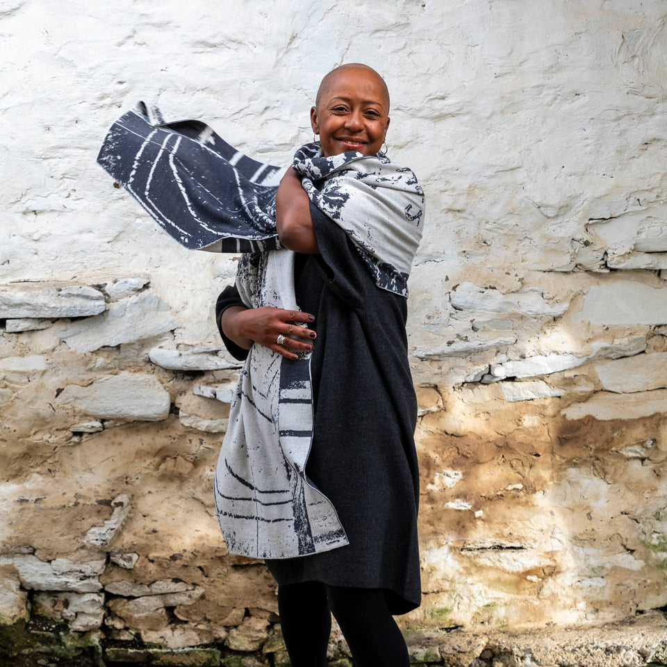 A black woman with a shaved head stands in a rustic stone building with whitewashed walls in Hoswick, Shetland. She wears black tights, a black charcoal woollen dress and wraps a large off white and charcoal shawl around herself, throwing one side over her shoulder. She smiles and looks at the camera,
