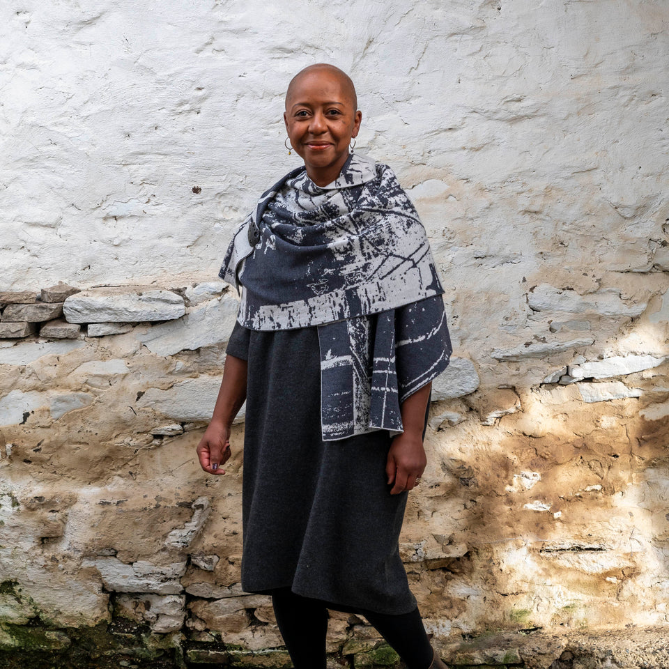 A black woman with a shaved head stands in a rustic stone building with whitewashed walls in Hoswick, Shetland. She wears black tights, a charcoal and off white shawl is wrapped around her.