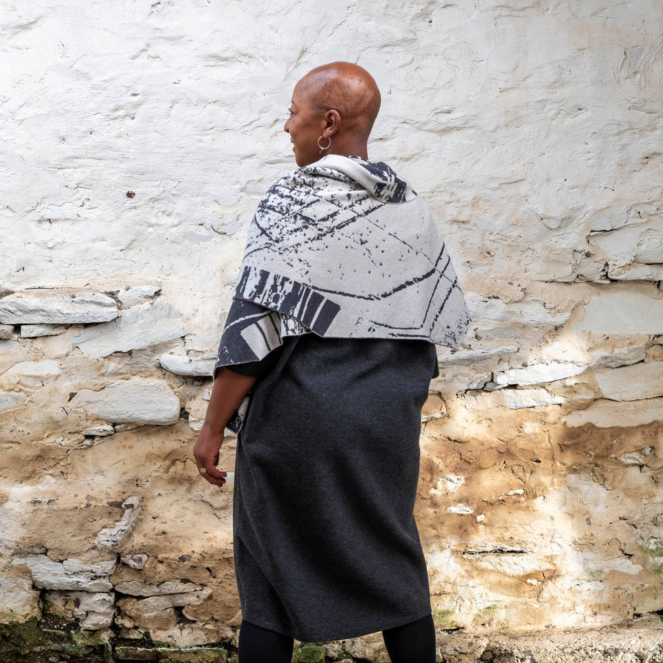 A black woman with a shaved head stands in a rustic stone building with whitewashed walls in Hoswick, Shetland. She wears black tights, a charcoal and off white shawl is wrapped around her. She has her back to the camera showing the angular edges of the shawl