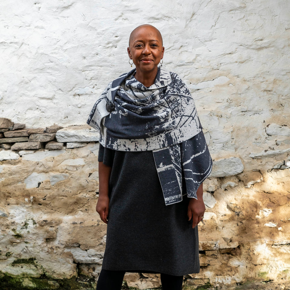 A black woman with a shaved head stands in a rustic stone building with whitewashed walls in Hoswick, Shetland. She wears black tights, a charcoal and off white shawl is wrapped around her.