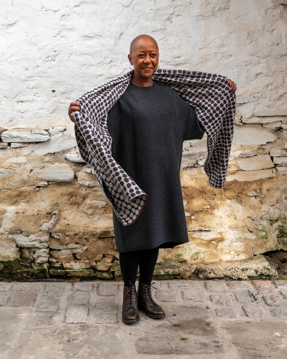 A black woman with a shaved head stands in a rustic stone building with whitewashed walls and a stone flat floor in Shetland. She wears a finely knitted modern shawl drapaed unfolded around her shoulders and down over her dress.. It is charcoal grey with pale grey irregular dots and a thin meandering line in cherry red. She also wears a charcoal woollen dress in a loose fit, black tights and dark brown brogue boots. She lifts the shawl around her neck and smiles