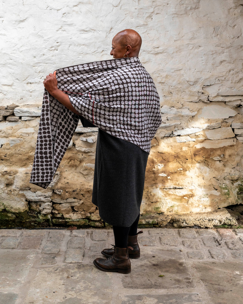 A black woman with a shaved head stands in a rustic stone building with whitewashed walls and a stone flat floor in Shetland. She wears a finely knitted modern shawl drapaed unfolded around her shoulders and down over her dress.. It is charcoal grey with pale grey irregular dots and a thin meandering line in cherry red. She also wears a charcoal woollen dress in a loose fit, black tights and dark brown brogue boots.