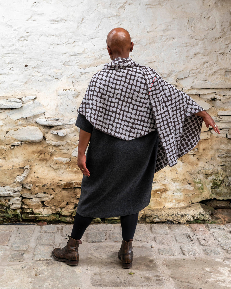 A black woman with a shaved head stands in a rustic stone building with whitewashed walls and a stone flat floor in Shetland. She wears a finely knitted modern shawl around her shoulders. It is charcoal grey with pale grey irregular dots and a thin meandering line in cherry red. She also wears a charcoal woollen dress in a loose fit, black tights and dark brown brogue boots. She stands with her back to the camera