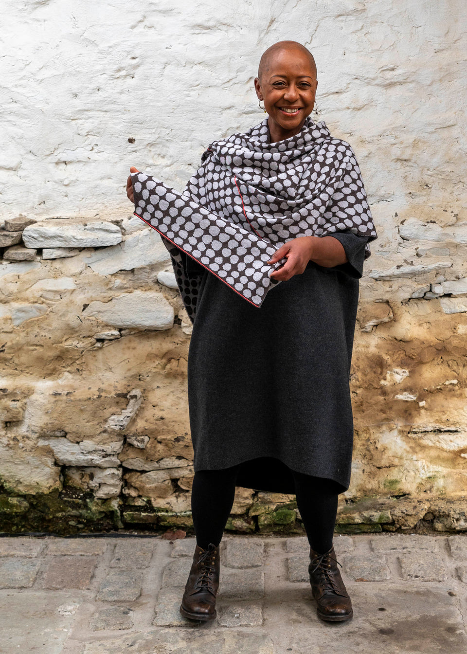 A black woman with a shaved head stands in a rustic stone building with whitewashed walls and a stone flat floor in Shetland.  She wears a finely knitted modern shawl around her shoulders. It is charcoal grey with pale grey irregular dots and a thin meandering line in cherry red. She also wears a charcoal woollen dress in a loose fit, black tights and dark brown brogue boots.