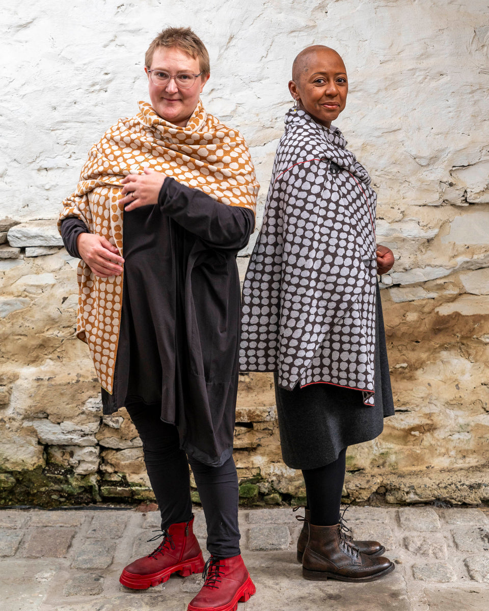 Two women stands in a whitewashed stone shed with flagstoned floor. On the left a white woman with fair hair wears a modern shawl in an ochre colour with irregular dots over a black tunic and leggings. On the right, a black woman with a shaved head wears the same shawl but in charcoal grey and pale grey over a grey woollen dress.