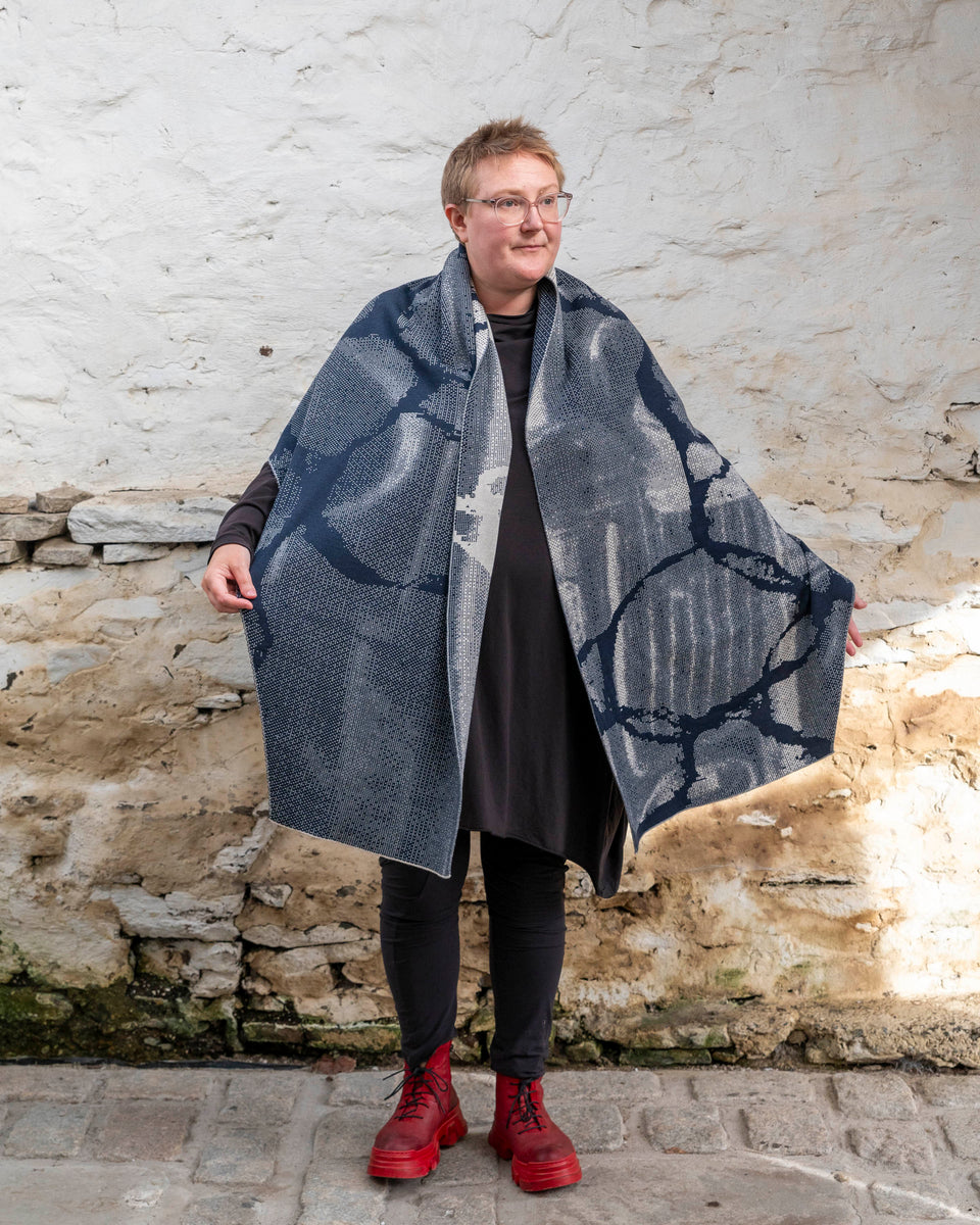 A white woman with short, fair hair stands inside a rustic stone building in Hoswick, Shetland. She wears an off-black tunic with matching leggings and red platform boots. She wears a navy and off white oversized shawl around her shoulders with both sides hanging down over her body. She holds the edges out slightly to show the pattern