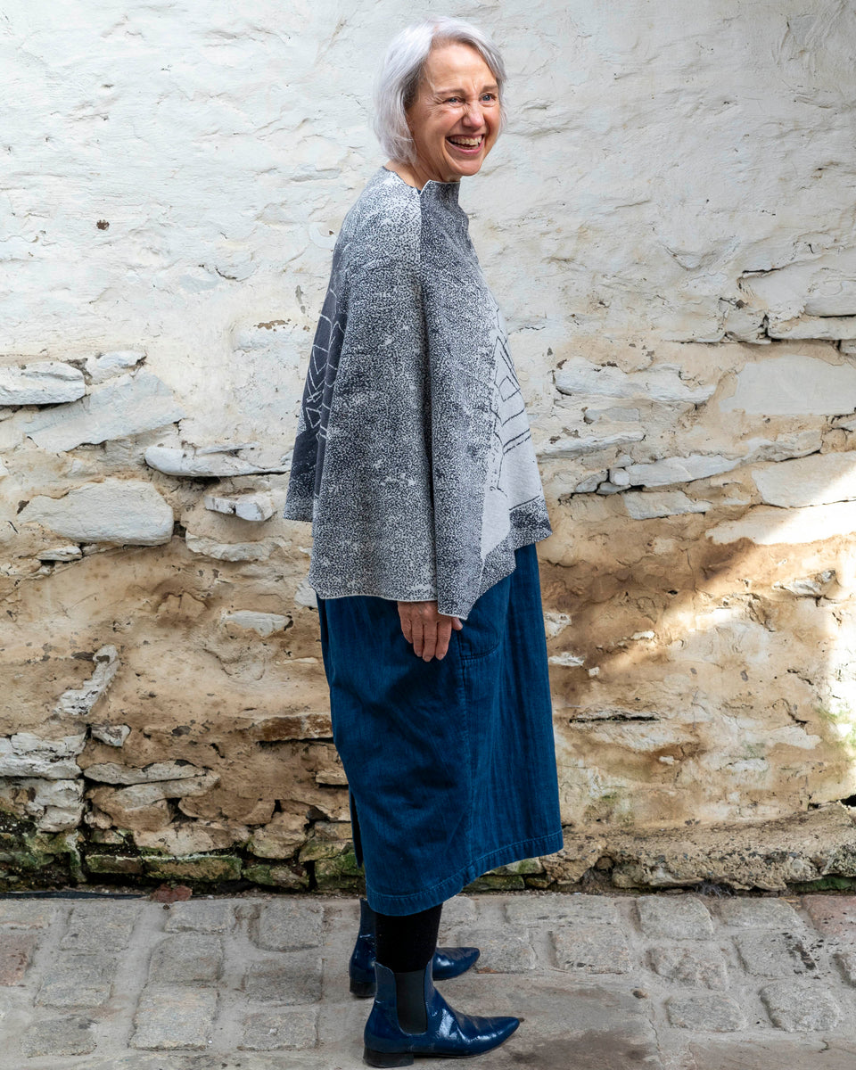 A woman with jaw length silver grey hair stands in a rustic, whitewashed stone building in Hoswick, Shetland. She is side on to the camera, she turns her head over her right shoulder and laughs . She is wearing a finely knitted piece of contemporary Scottish knitwear - a cape in charcoal and soft white. underneath she wears a navy t-shirt and a loose indigo denim dress. She wears patent navy pointy dealer boots