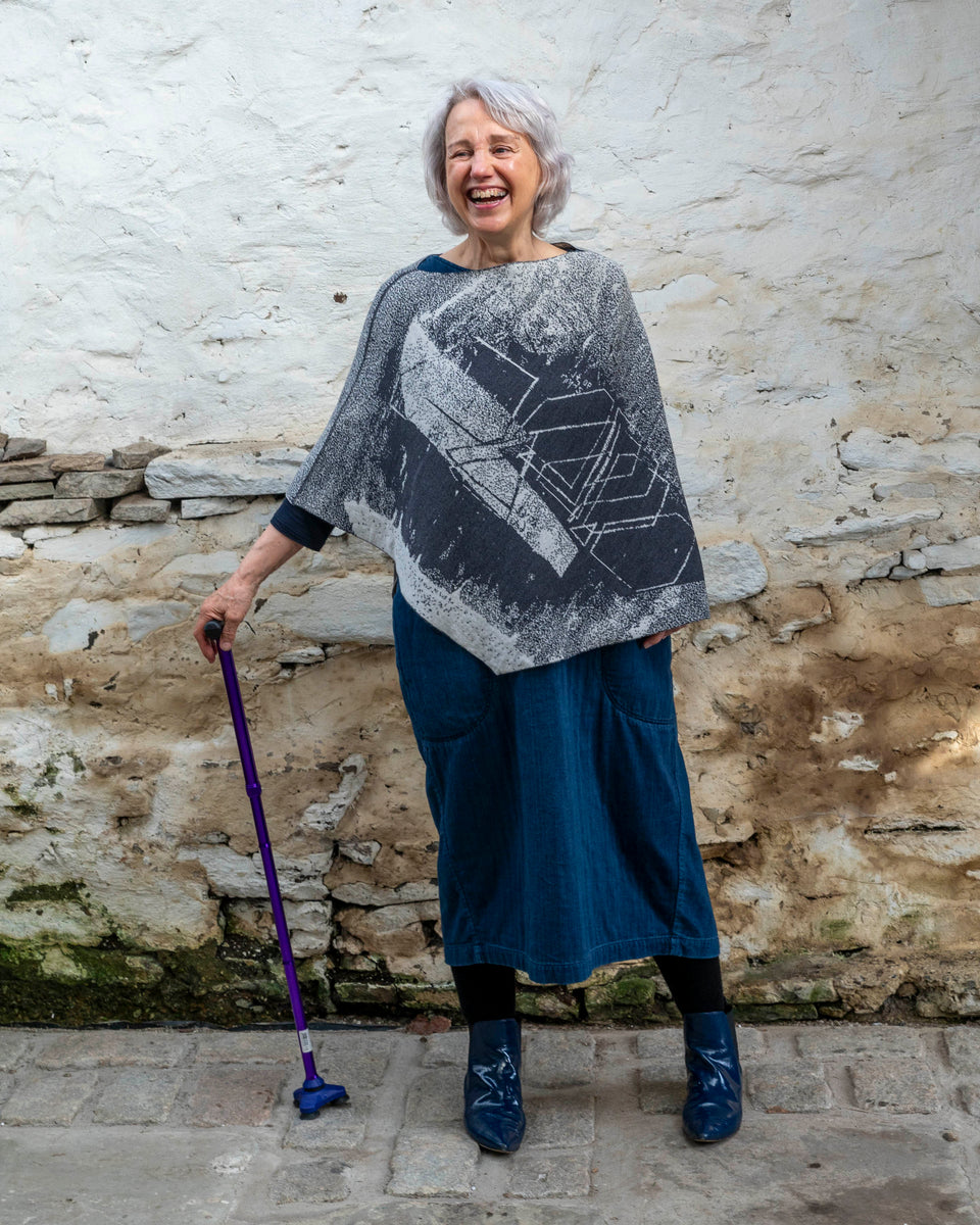 A woman with jaw length silver grey hair stands in a rustic, whitewashed stone building in Hoswick, Shetland. She holds a purple walking stick with her right hand and laughs, looking to her right. She is wearing a finely knitted piece of contemporary Scottish knitwear - a cape in charcoal and soft white. underneath she wears a navy t-shirt and a loose indigo denim dress. She wears patent navy pointy dealer boots