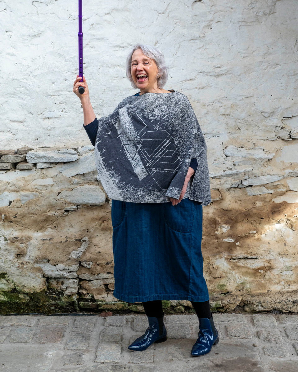 A woman with jaw length silver grey hair stands in a rustic, whitewashed stone building in Hoswick, Shetland. She holds a purple walking stick up in the air with right right hand and laughs. She is wearing a finely knitted piece of contemporary Scottish knitwear - a cape in charcoal and soft white. underneath she wears a navy t-shirt and a loose indigo denim dress. She wears patent navy pointy dealer boots.