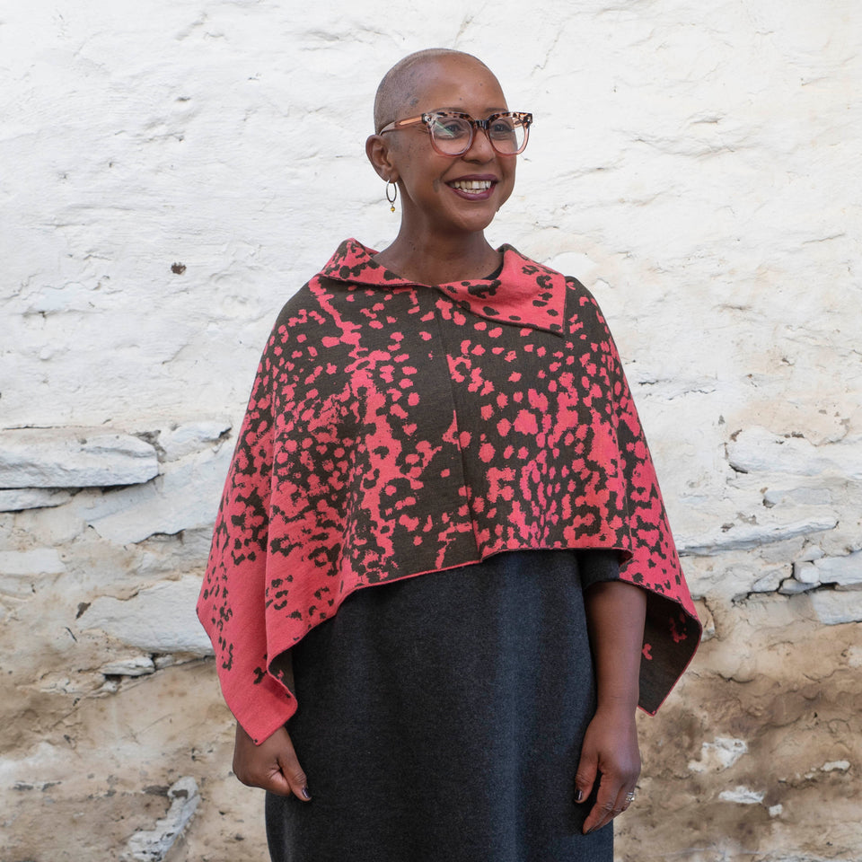 A black woman with a shaved wears tortoiseshell glasses and hooped silver earrings. She looks slightly away from the camera and smiles. She is wearing a knitted cape with an abstract speckled pattern in pink and dark green over a charcoal grey dress. She's standing in a rustic, stone, whitewashed Shetland shed.