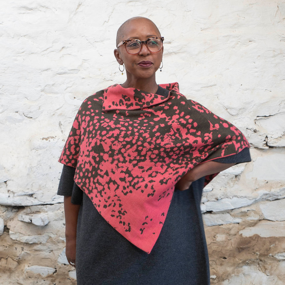 A black woman with a shaved wears tortoiseshell glasses and hooped silver earrings. She looks slightly away from the camera and pouts. She is wearing a knitted cape with an abstract speckled pattern in pink and dark green over a charcoal grey dress. She's standing in a rustic, stone, whitewashed Shetland shed.