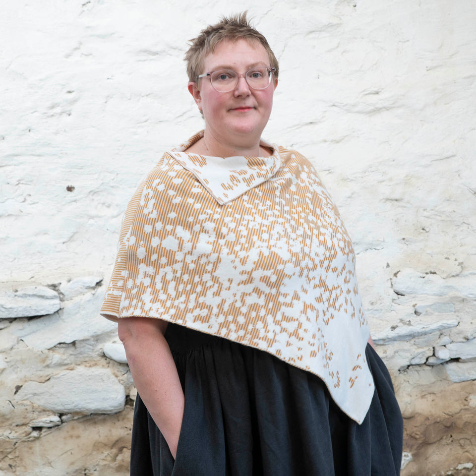 Felix Ford stands in a rustic, whitewashed stone shed in Hoswick, Shetland. A white woman with short, fair hair wears spectacles and - over a black linen dress with gathered skirt - a finely knitted cape in a speckled pattern in cream, gold and brown. She looks towards the camera with a steady gaze and has her hands in the pockets of her dress.