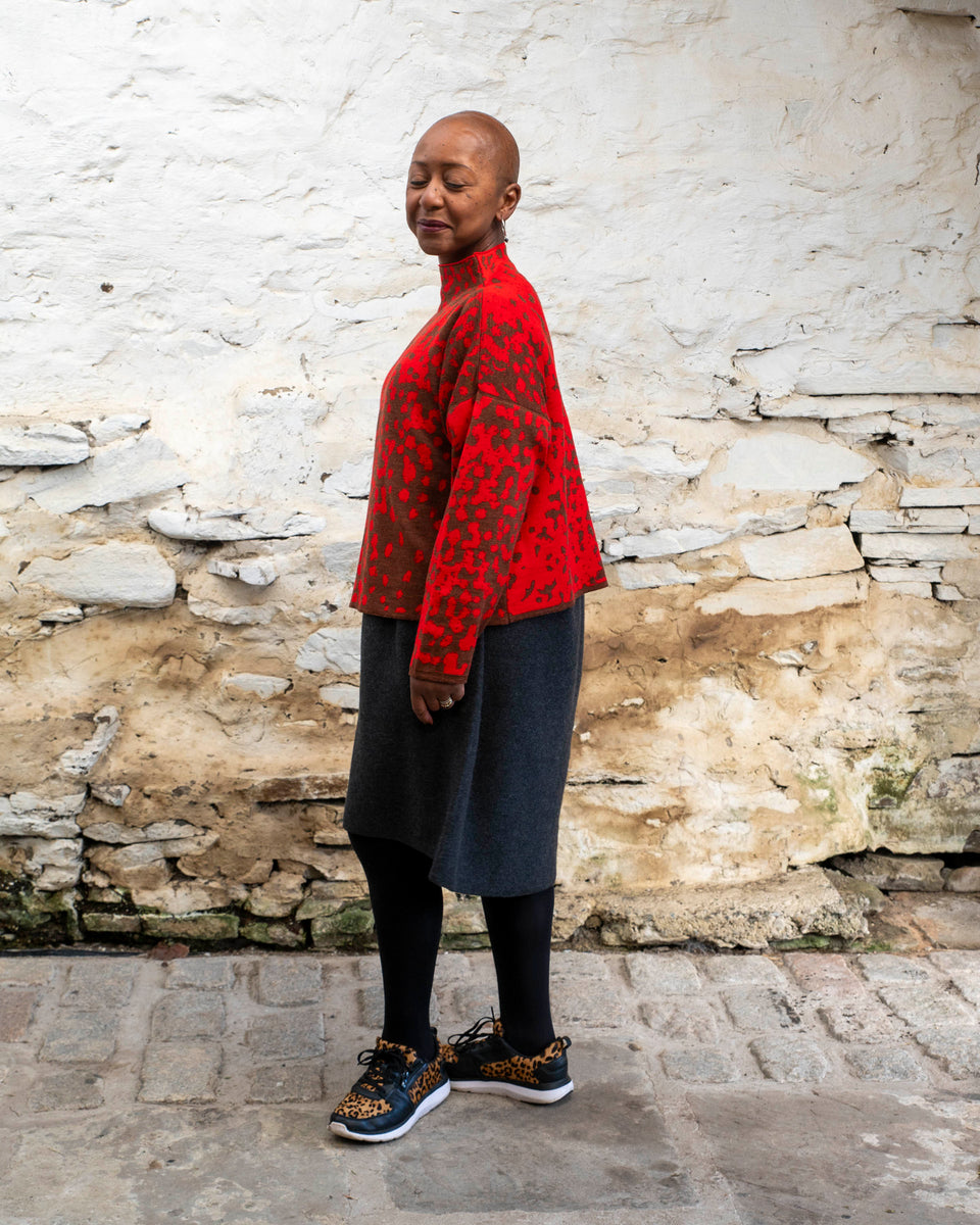 Jeanette Sloan, a black woman with a shaved head stands inside a rustic, whitewashed, stone building in Hoswick Shetland. She is wearing largish, silver hooped earrings with a bead hanging, a tomato red and chestnut brown finely knitted boxy jumper with a graphic animal print patter and a high neck, a carcoal grey woolen dress, black opaque tights and black leather and leopard print trainers. She is standing side on and closes her eyes as she smiles.