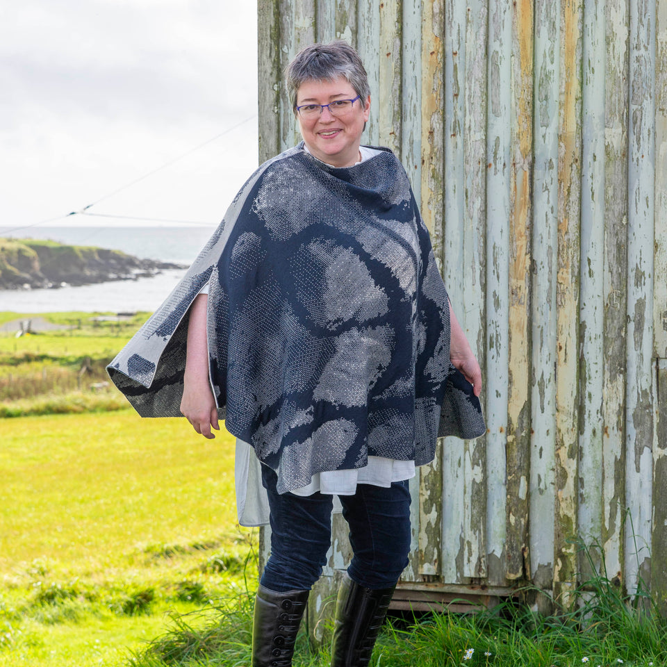 A white woman with short brown and grey hair stands in front of an old timber building with peeling paint in Hoswick, Shetland. She is wearing purple, metal rimmed glasses, navy velvet leggings tucked into black leather knee high boots and a finely knitted navy and off white cape in an abstract pattern. Underneath is a mens white, dress shirt. In the background is Hoswick bay.