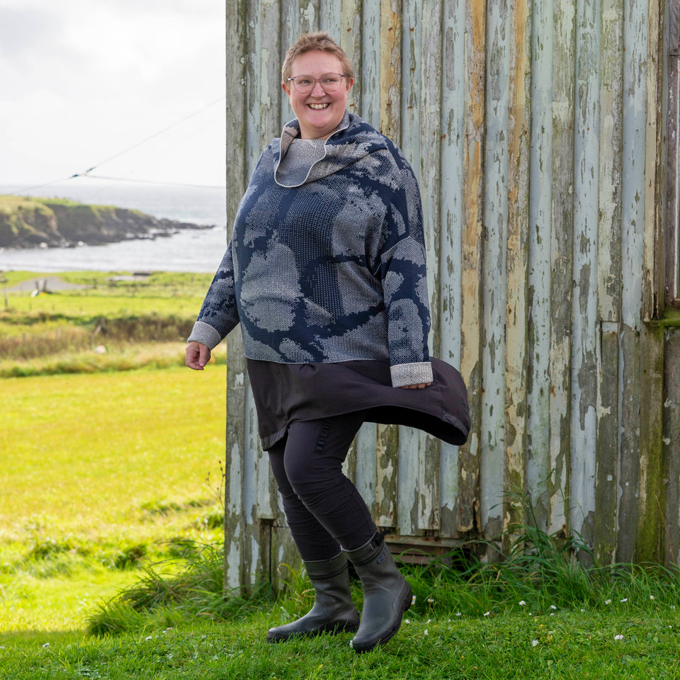 Felix Ford wears a Nielanell cowl-necked jumper finely knitted in dark navy and off white. Abstract patterns form larger motifs. Felix stands in front of the Gospel Hall, Hoswick with the sea behind.