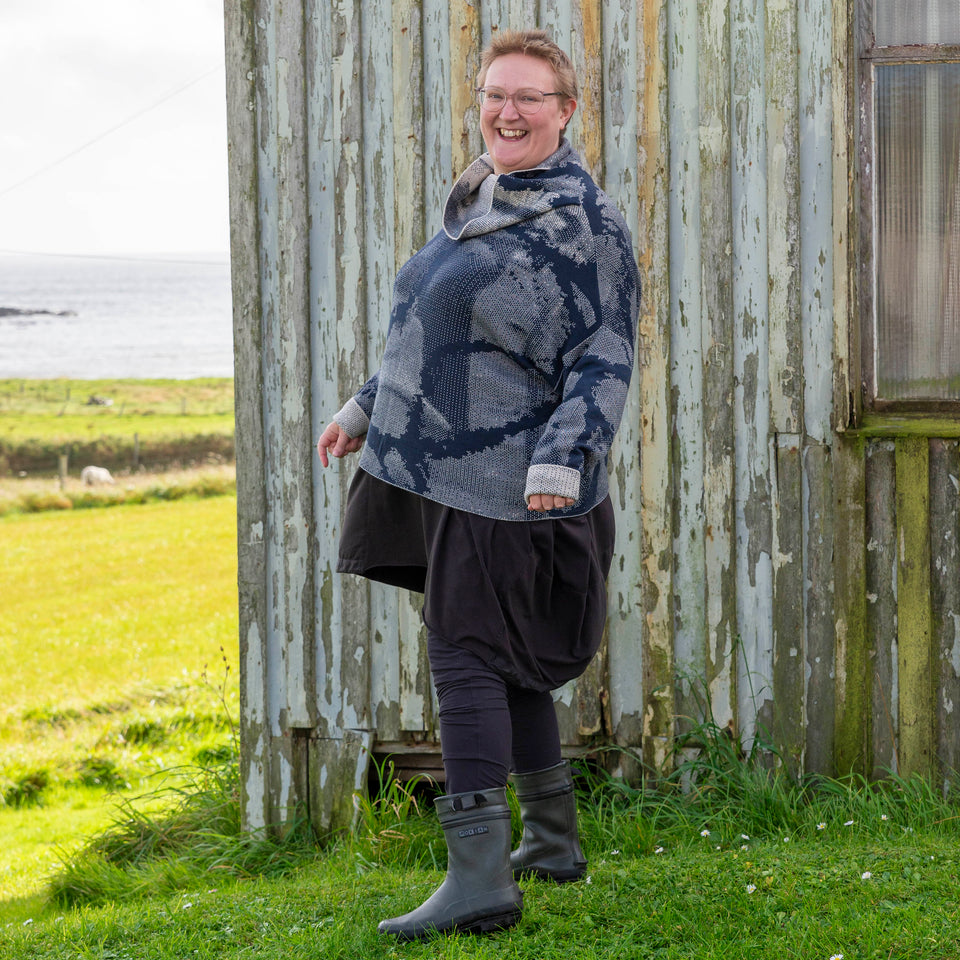 A white woman with short fair hair stands in front of the wooden Gospel Hall at Hoswick, shetland. She is wearing a finely knitted Shetland jumper in dark navy and off white. It has a cowl neck and abstract patterning. She also wears an off-black tunic, leggings and short green wellies. The sea is in the background. She stands with her left shoulder dropped towards the camera and smiles.