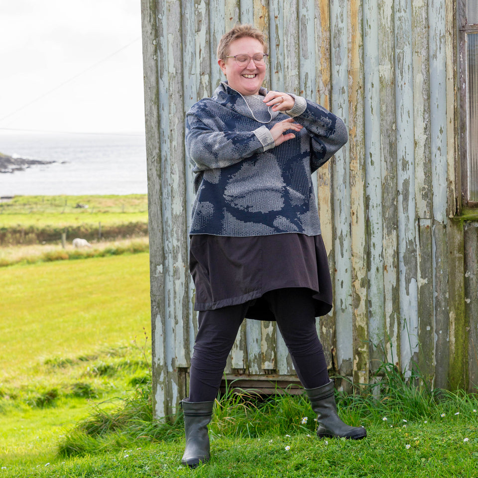A white woman with short fair hair stands in front of the wooden Gospel Hall at Hoswick, shetland. She is wearing a finely knitted Shetland jumper in dark navy and off white. It has a cowl neck and abstract patterning. She also wears an off-black tunic, leggings and short green wellies. The sea is in the background. She adjusts the turn-backs at the jumper cuffs and smiles.