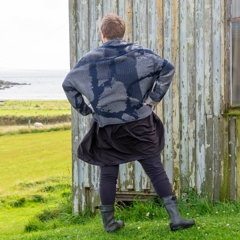 A white woman with short fair hair stands in front of the wooden Gospel Hall at Hoswick, shetland. She is wearing a finely knitted Shetland jumper in dark navy and off white. It has a cowl neck and abstract patterning. She also wears an off-black tunic, leggings and short green wellies. The sea is in the background. She stands with her back to the camera with her hands on her hips.