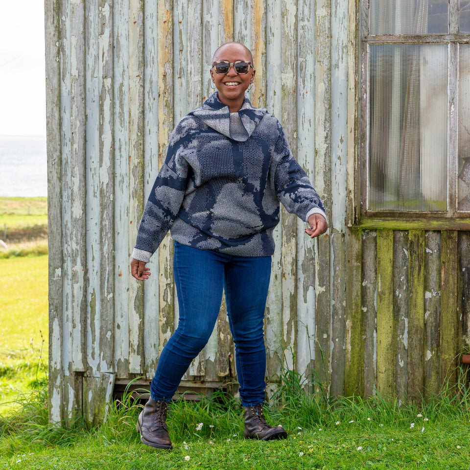 Jeanette Sloan at Hoswick, Shetland. She stands on the grass with sea behind. She wears tortoiseshell sunglasses, hoop earrings, a cowl necked Nielanell jumper in navy and off white abstract pattern, dark indigo jeans and brown lace up boots.