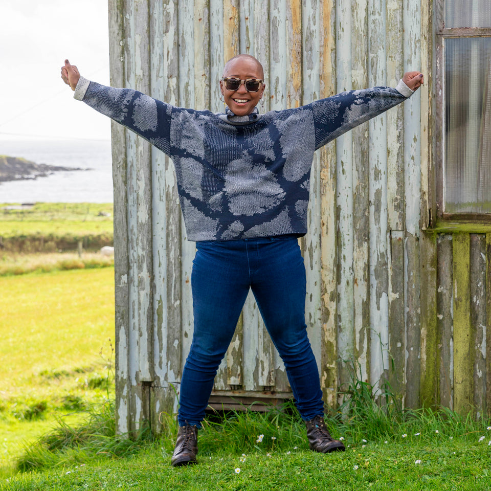 Jeanette Sloan at Hoswick, Shetland. She stands on the grass with sea behind. She wears tortoiseshell sunglasses, hoop earrings, a cowl necked Nielanell jumper in navy and off white abstract pattern, dark indigo jeans and brown lace up boots.
