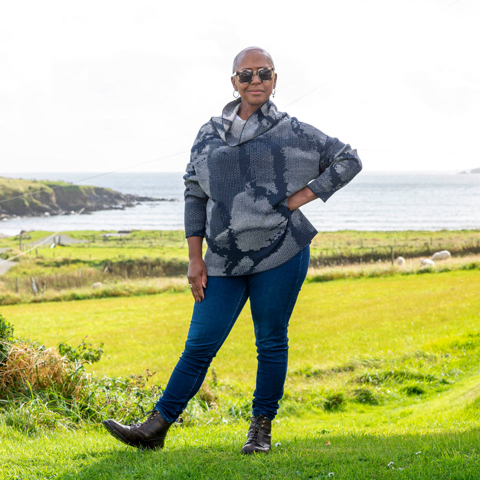 Jeanette Sloan at Hoswick, Shetland. She stands on the grass with sea behind. She wears tortoiseshell sunglasses, hoop earrings, a cowl necked Nielanell jumper in navy and off white abstract pattern, dark indigo jeans and brown lace up boots. Her right hand rests at her waist.