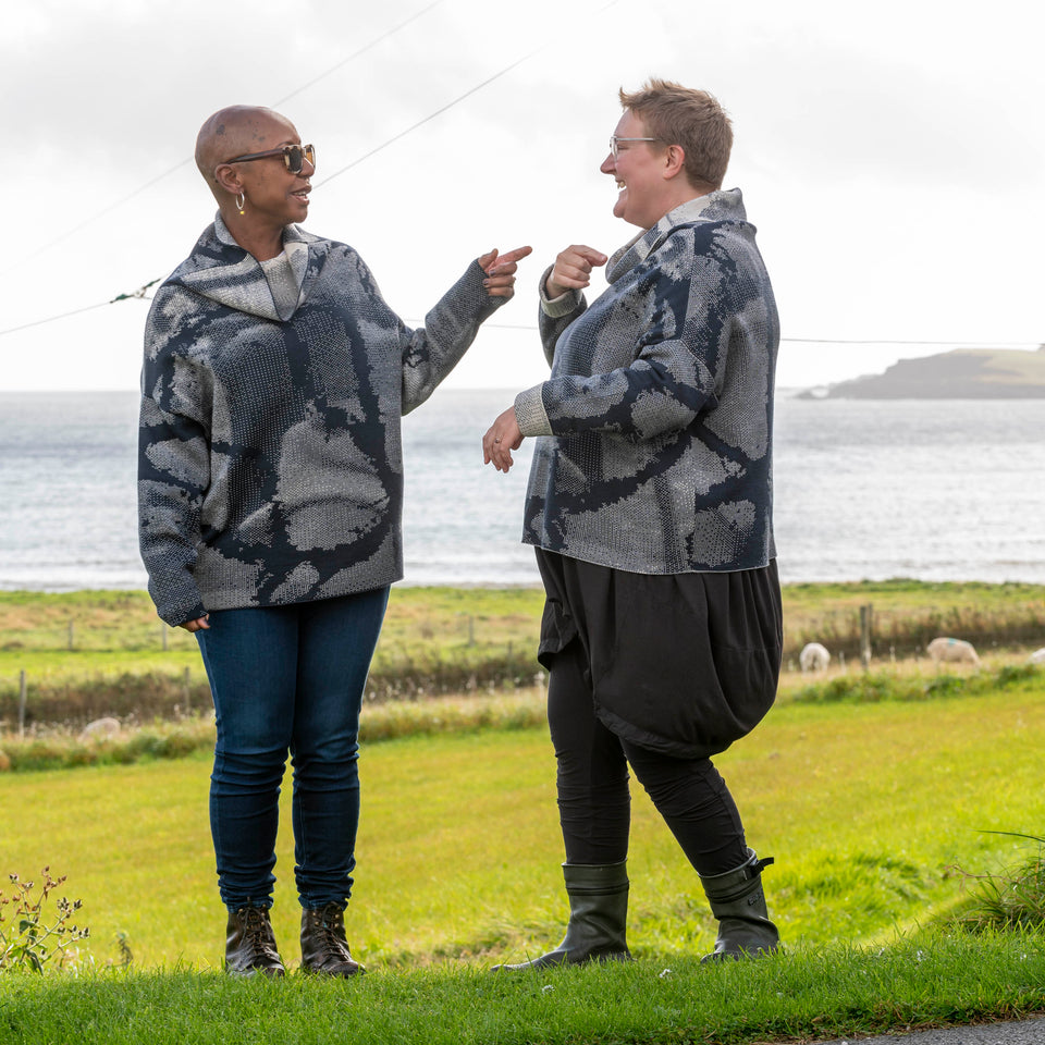 One black and one white woman on the grass at Hoswick, Shetland with the sea behind. They are both wearing the same cowl-necked contemporary merino jumper. A contemporary Shetland sweater in navy and off white with an abstract pattern.