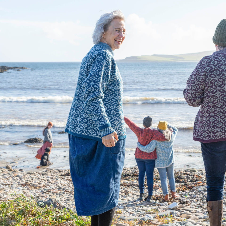 A woman with jaw length grey hair wears a blue and grey fair isle patterned jumper over a loose denim dress. She turns to face the camera. In the background, the sea at Hoswick stretches out towards Levenwick headland. On the shore a young woman with a black labrador and tow women with a rusty fair isle jumper and a pale blue and pale grey fair isle jumper are milling about. All three on the beach are wearing beanie hats.